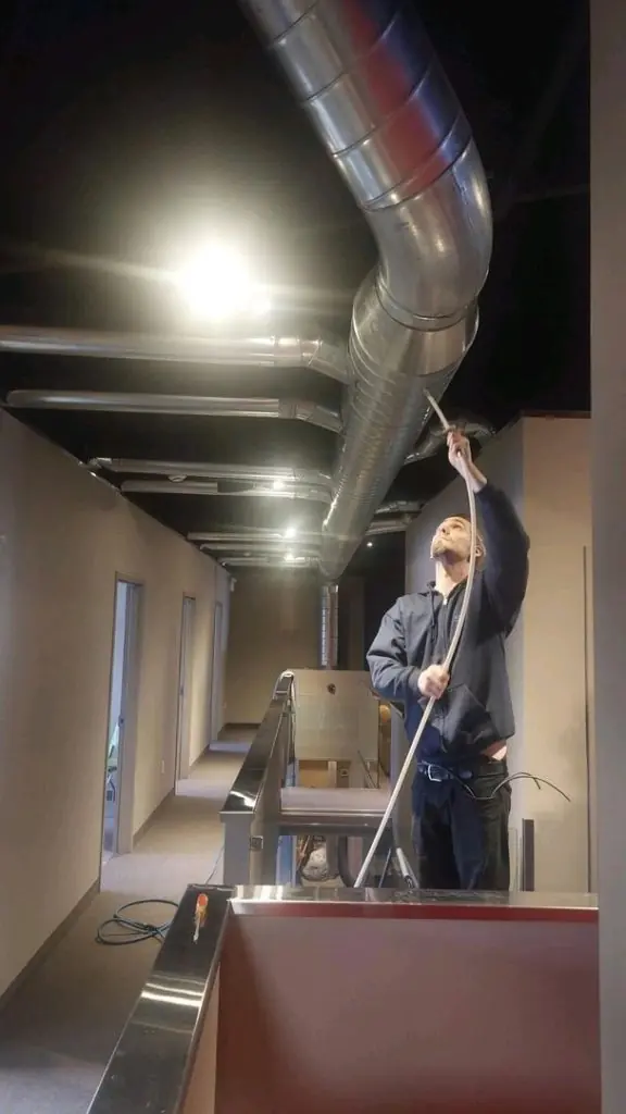 A worker cleaning large metallic air ducts on the ceiling of an office building using a long tool.