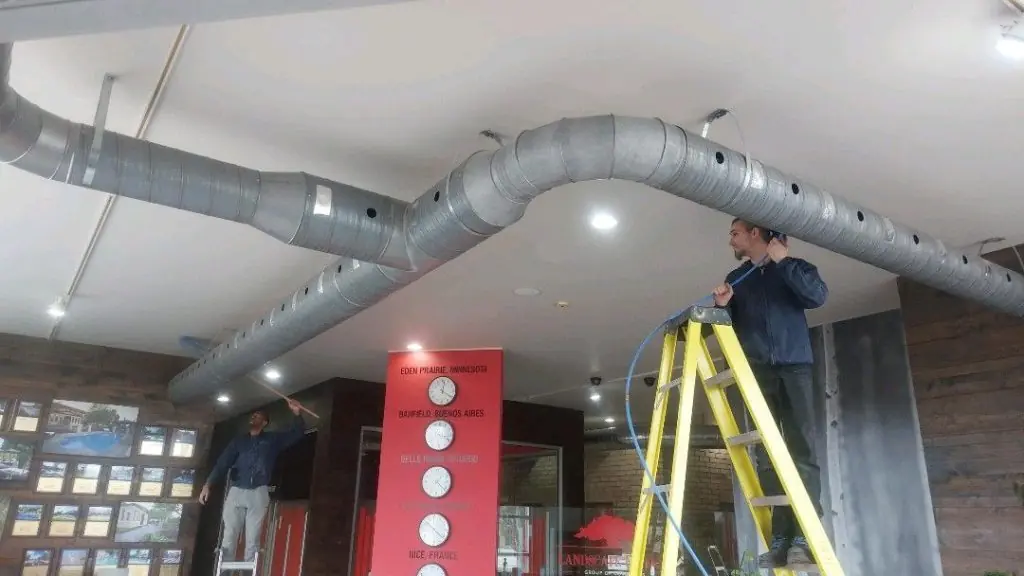 Two technicians working on an overhead duct system in a commercial space, one on a ladder and one reaching up from the floor.