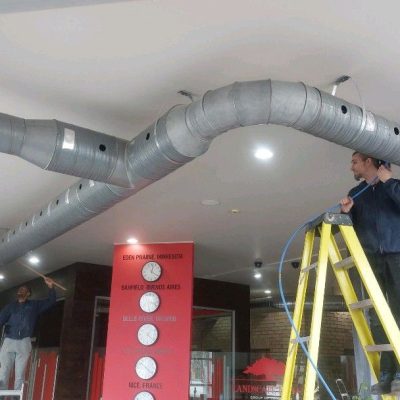 Two technicians working on an overhead duct system in a commercial space, one on a ladder and one reaching up from the floor.